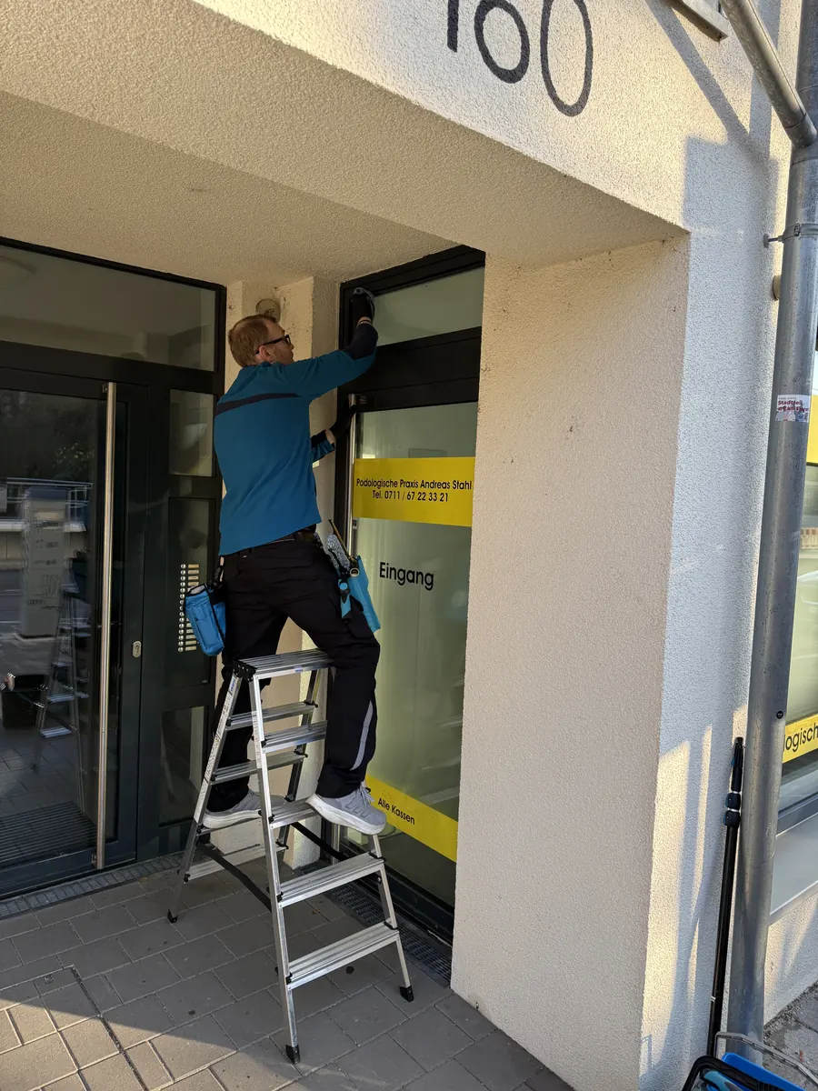 Praxisreinigung: Mitarbeiter von H2 Clean reinigt auf der Leiter Fensterrahmen und Eingangsbereich einer Podologiepraxis